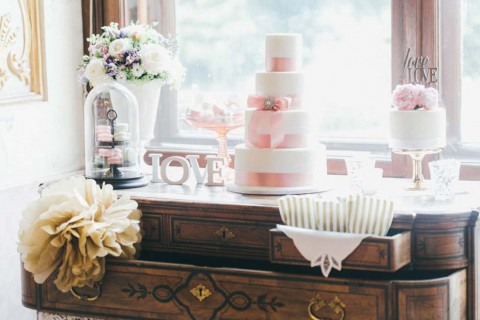 Hochzeitswahn - Eleganter Hochzeitsdesserttisch mit einer großen, dreistöckigen rosa-weißen Torte, die mit einer Schleife geschmückt ist, kleineren Torten und einem Blumenarrangement, aufgestellt neben einem Fenster mit einem „Love“-Schild. Schloss-Eckartsau