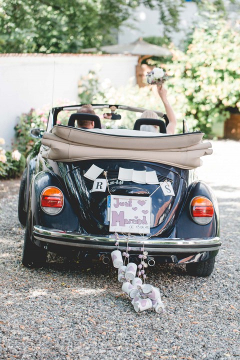 Hochzeitswahn - Ein frisch verheiratetes Paar fährt in einem Oldtimer-Cabrio los, das mit einem „Just Married“-Schild geschmückt ist und hinter dem Dosen hängen. Die Braut wirft freudig ihren Blumenstrauß vom Rücksitz aus in die Luft.