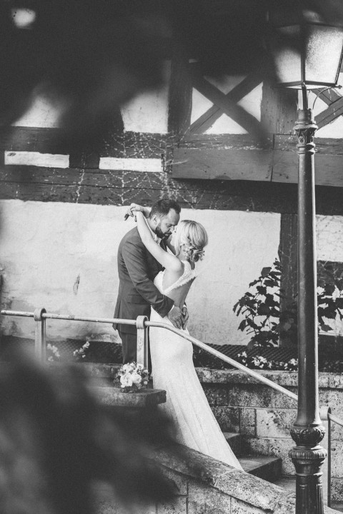 Hochzeitswahn - Ein monochromes Bild von Braut und Bräutigam, die sich auf einer Steintreppe einen romantischen Kuss geben, eingerahmt von einer Holzbalkenkonstruktion und einem Metallgeländer, mit sanfter Beleuchtung durch eine Laterne darüber.