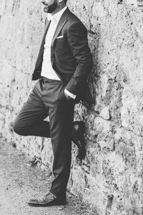 Hochzeitswahn - Ein Schwarzweißfoto eines Mannes im Anzug, der an einer strukturierten Steinwand lehnt. Er ist teilweise abgewandt, mit einem Fuß gegen die Wand gestützt, und strahlt eine entspannte und dennoch stilvolle Haltung aus.