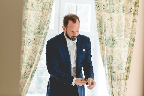 Hochzeitswahn - Ein Mann im blauen Anzug richtet seine Manschettenknöpfe an einem Fenster, durch dessen mit Dollarscheinen gemusterte Vorhänge Sonnenlicht fällt. Er trägt einen Bart und ist auf seine Aufgabe konzentriert.