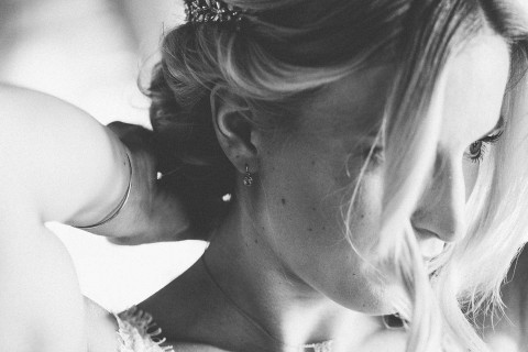 Hochzeitswahn - Schwarzweißfoto einer Nahaufnahme einer Frau mit Fokus auf ihr Profil. Sie trägt eine elegante Frisur mit Haarschmuck und einem zarten Ohrring und man sieht einen Hauch ihres Spitzenkleides. Die Beleuchtung hebt ihre Gesichtszüge sanft hervor.