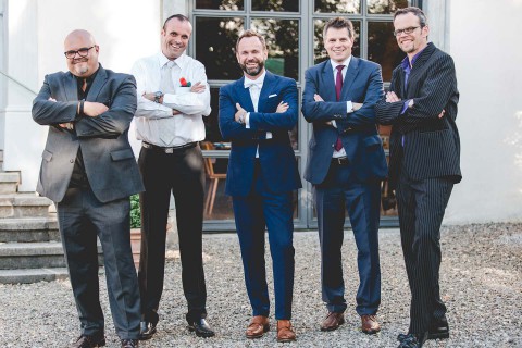 Hochzeitswahn - Fünf Männer in Geschäftskleidung stehen mit verschränkten Armen vor einem Gebäude und lächeln in die Kamera. Zwei tragen eine Brille und einer hat eine rote Ansteckblume an seiner Jacke. Sie zeigen ein selbstbewusstes, professionelles Auftreten.