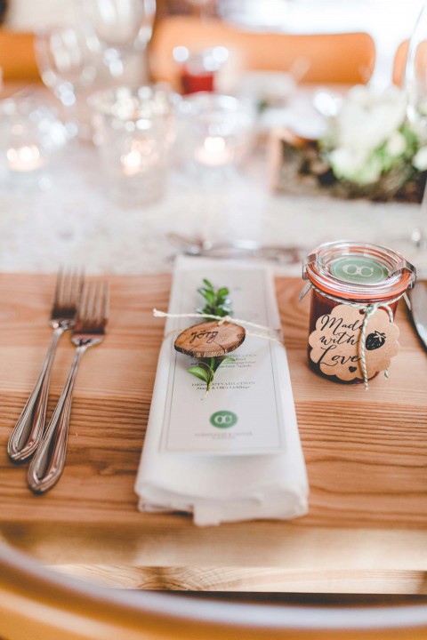 Hochzeitswahn - Ein Hochzeitsgedeck auf einem Holztisch mit einer hübsch arrangierten Serviette mit einer Menükarte, einem kleinen hölzernen Namensschild und Besteck. Ein Glasgefäß mit der Aufschrift „Danke“ und ein Pflanzenzweig akzentuieren das Arrangement.