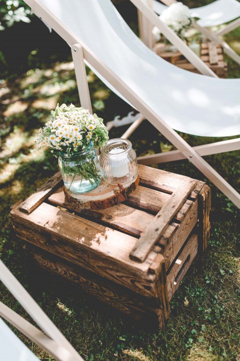 Hochzeitswahn - Eine rustikale Holzkiste dient als Tisch und enthält eine Glasvase mit Wildblumen und ein Einmachglas. Sie steht auf einer Rasenfläche mit weißen Liegestühlen im Hintergrund.