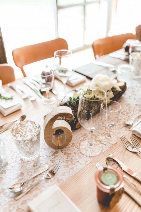 Hochzeitswahn - Elegant gedeckter Esstisch für ein formelles Essen mit strahlend weißem Geschirr, mehreren Gläsern und Besteck auf einer gemusterten Tischdecke. Ein hölzernes Tafelaufsatz mit Blumen und Grünpflanzen verläuft entlang des Tisches und wird von sanftem, natürlichem Licht erhellt.