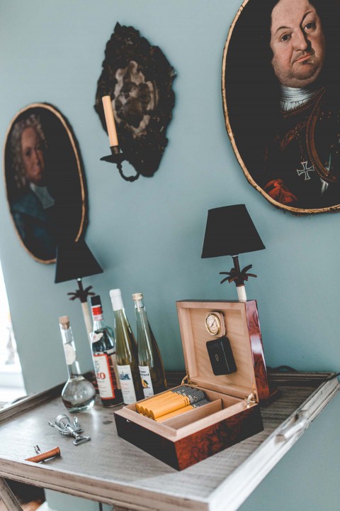 Hochzeitswahn - Eine elegante Zimmerecke im Vintage-Stil mit einem Tisch mit einer offenen Zigarrenkiste, verschiedenen Schnapsflaschen und einer kleinen Lampe. Porträts in verzierten Rahmen schmücken die blaugrüne Wand und schaffen ein historisches Ambiente.