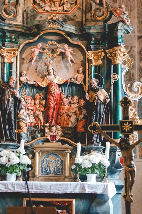 Hochzeitswahn - Ein kunstvoller Kirchenaltar mit einer zentralen Statue der Jungfrau Maria, umgeben von verschiedenen religiösen Figuren, geschmückt mit komplizierten goldenen Elementen, Blumenarrangements und flackernden Kerzen.