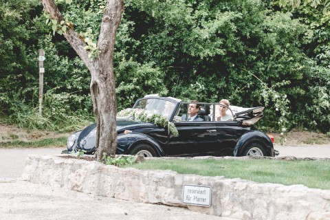 Hochzeitswahn - Ein frisch verheiratetes Paar fährt in einem mit Blumen geschmückten schwarzen Oldtimer-Cabrio durch eine parkähnliche Umgebung mit grünen Büschen und Bäumen. Im Vordergrund ist ein Schild mit der Aufschrift „Reserviert für Partner“ zu sehen.