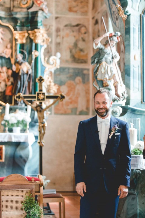 Hochzeitswahn - Ein lächelnder Mann in einem dunkelblauen Anzug mit einer Ansteckblume steht in einer Kirche, umgeben von kunstvollen religiösen Fresken und Goldverzierungen. Sein Gesichtsausdruck ist fröhlich und die Kulisse reich an Geschichte.