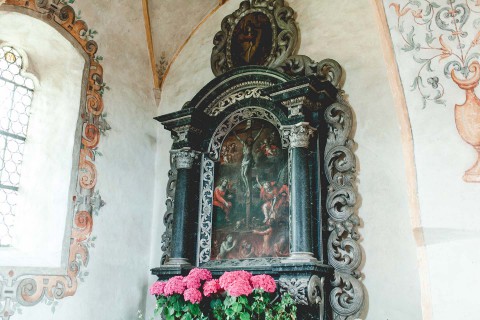 Hochzeitswahn - Ein reich verzierter religiöser Schrein in einem Raum mit Fresken an den Wänden. Der Schrein zeigt eine detailreiche Skulptur mit Figuren, eingerahmt von einem kunstvollen schwarz-goldenen Bogen über einer Ausstellung rosa Hortensien.
