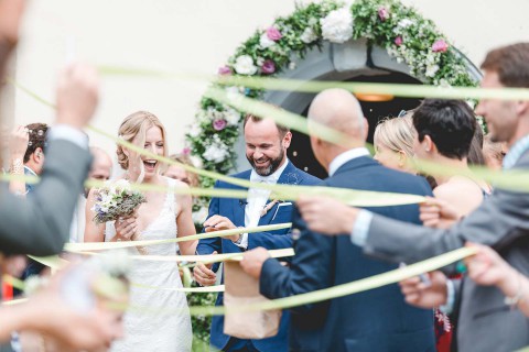 Hochzeitswahn - Ein fröhliches Brautpaar im Hochzeitskleid schreitet, umschlungen von Bändern, die die Gäste halten, durch eine fröhliche Menschenmenge unter einem dekorativen Torbogen hindurch.