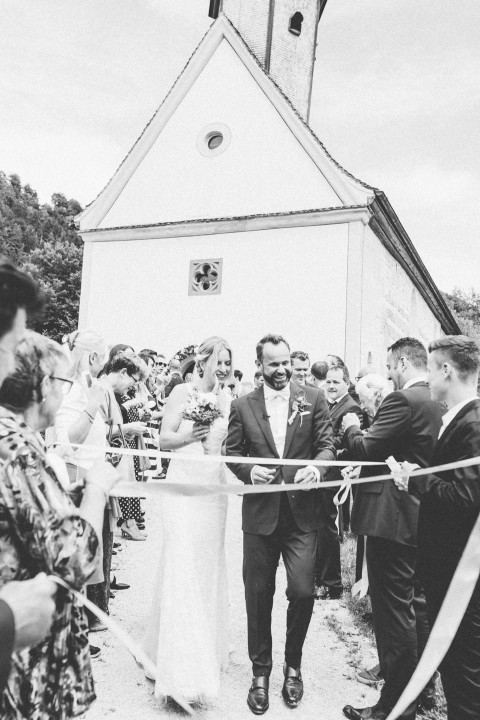 Hochzeitswahn - Schwarzweißfoto eines fröhlichen Brautpaares, das durch eine Menschenmenge vor einer Kirche geht, während die Gäste feiernd Bänder über ihren Weg halten.