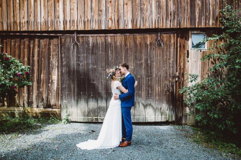 Hochzeitswahn - Braut und Bräutigam küssen sich vor einer rustikalen Scheune mit verwitterten Holzwänden. Die Braut trägt ein langes weißes Kleid und hält einen Blumenstrauß, während der Bräutigam einen blauen Anzug und braune Schuhe trägt.