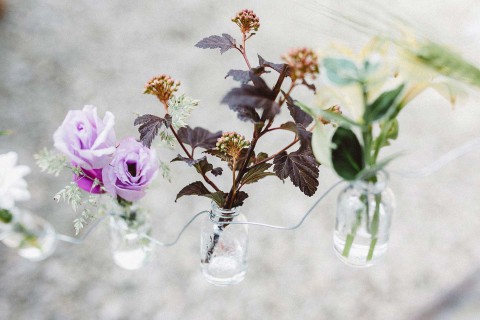 Hochzeitswahn - Drei kleine, durch eine Schnur verbundene Glasgefäße, in denen sich jeweils verschiedene Wildblumen befinden, darunter eine violette Rose. Sie stehen auf einer rauen Betonoberfläche und bilden so einen rustikalen und zarten Kontrast.