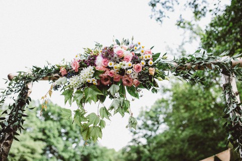 Hochzeitswahn - Ein wunderschön dekorierter Blumenbogen mit leuchtend rosa, weißen und gelben Blumen, durchsetzt mit üppigen grünen Blättern, vor einem sanft verschwommenen Hintergrund aus Bäumen.