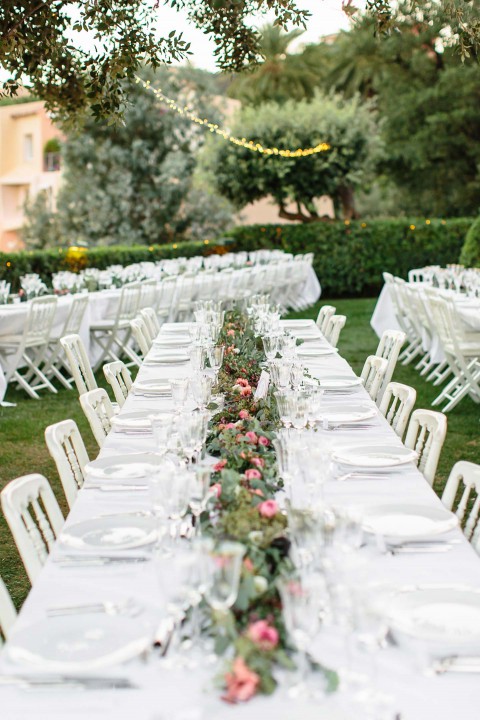 Hochzeitswahn - Ein elegant gedeckter Esstisch im Freien mit weißen Stühlen, feinem Porzellan und einem Blumenschmuck in der Mitte. Lichterketten und üppige grüne Bäume verstärken die Atmosphäre im Hintergrund.