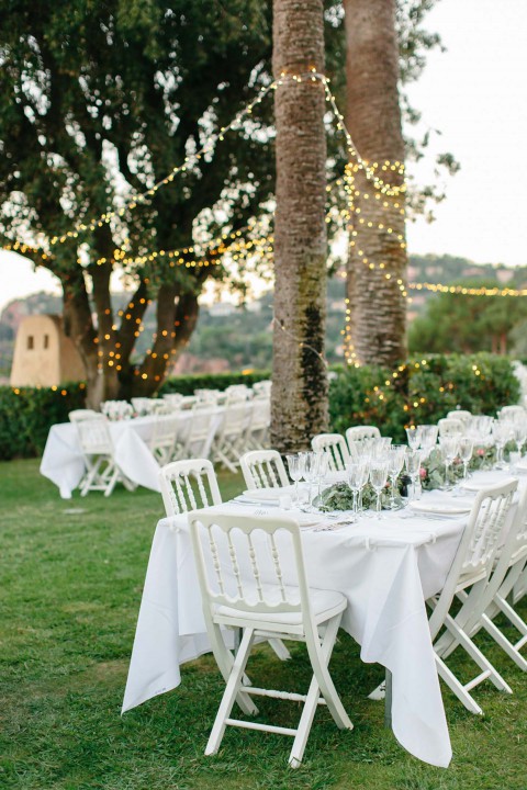 Hochzeitswahn - Empfangsbereich für eine Hochzeit im Freien in der Abenddämmerung mit weißen Stühlen und Tischen, die mit Gläsern und Tellern gedeckt sind. Lichterketten sind um die nahegelegenen Palmen gewickelt und erzeugen ein warmes, einladendes Licht.