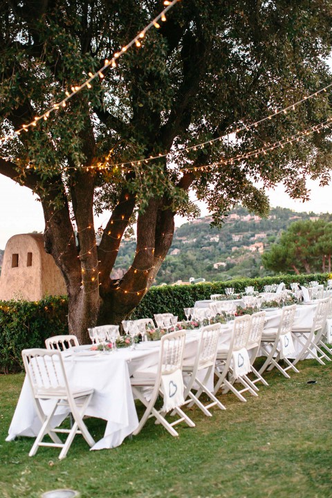 Hochzeitswahn - Ein Essbereich im Freien unter einem großen Baum mit Lichterketten, mit einem langen Tisch mit weißen Stühlen und elegantem Gedeck vor der Kulisse einer sanften Hügellandschaft.