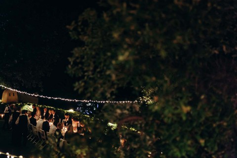 Hochzeitswahn - Eine abendliche Gartenparty mit Gästen, die an Tischen sitzen, die von Lichterketten beleuchtet werden und von üppigem grünem Laub umgeben sind. Die Szene vermittelt eine warme, intime Atmosphäre unter einem dunklen Himmel.
