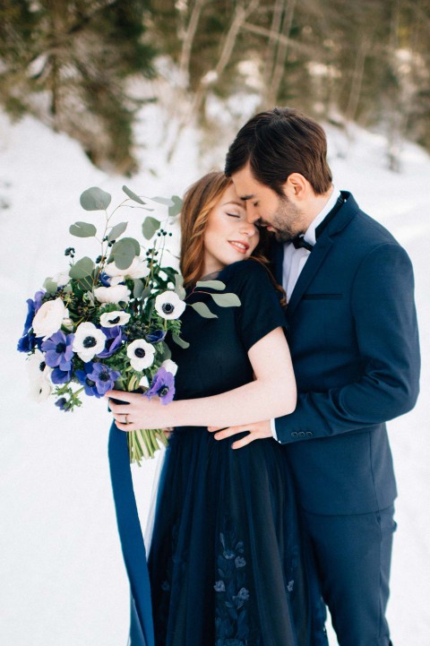 Hochzeitswahn - Ein Paar umarmt sich in einer verschneiten Landschaft. Die Frau im dunklen Kleid hält einen Strauß mit blauen und weißen Blumen, der Mann im blauen Anzug lehnt sanft seinen Kopf an ihren. Beide haben die Augen geschlossen, was einen heiteren Moment vermittelt.