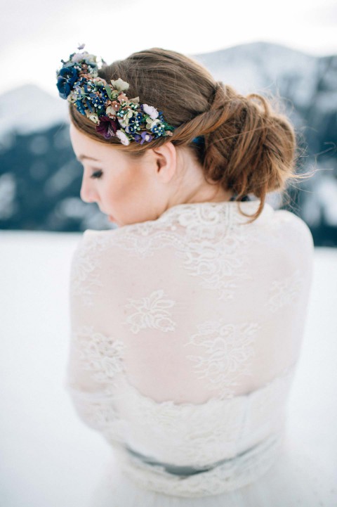 Hochzeitswahn - Eine Frau in einem Spitzenbrautkleid und einem bunten Blumenstirnband schaut weg, ihr Haar ist zu einem Dutt hochgesteckt. Im Hintergrund stehen schneebedeckte Berge.