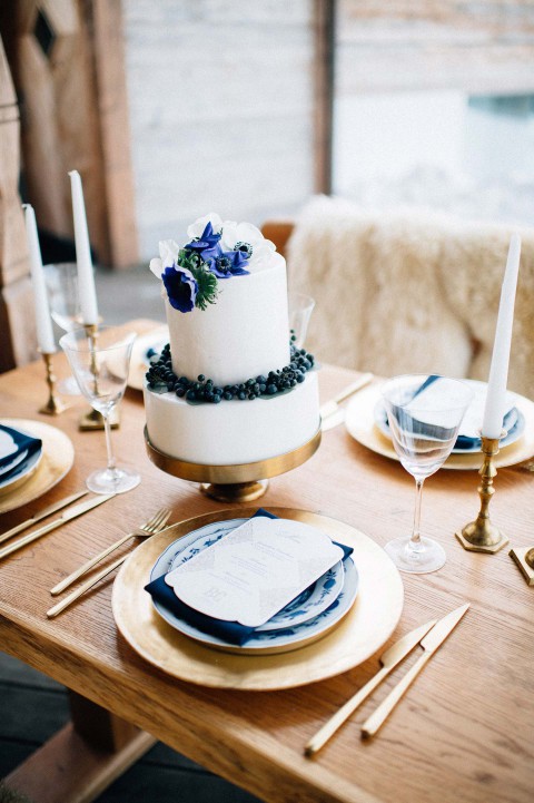 Hochzeitswahn - Eine dreistöckige Hochzeitstorte, geschmückt mit blauen und violetten Blumen und Brombeeren auf einem Holztisch. Elegante Gedecke mit blauen Tellern, goldenem Besteck und Kristallgläsern runden die Szene ab.