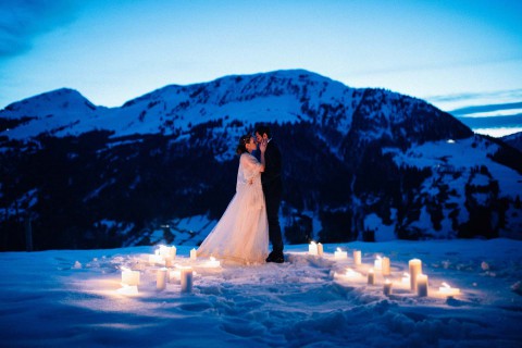 Hochzeitswahn - Braut und Bräutigam umarmen sich umgeben von brennenden Kerzen auf Schnee und im Hintergrund die Berge in der Abenddämmerung, was eine romantische, heitere Atmosphäre schafft.