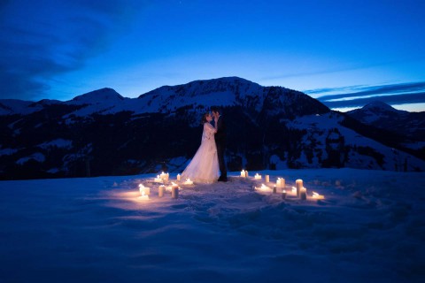 Hochzeitswahn - Ein Paar im Hochzeitskleid umarmt sich umgeben von Kerzen im Schnee in der Dämmerung, mit majestätischen schneebedeckten Bergen unter einem tiefblauen Himmel im Hintergrund.