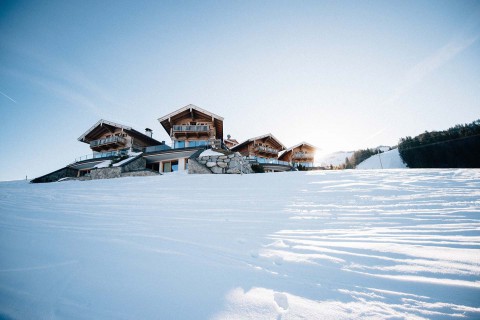 Hochzeitswahn - Eine malerische Winterlandschaft mit mehreren Holzchalets mit schneebedeckten Dächern, eingebettet in einen Hügel mit unberührtem Schnee im Vordergrund und einem klaren blauen Himmel.