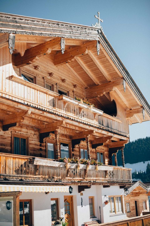 Hochzeitswahn - Ein traditionelles alpines Holzchalet mit aufwendigen Schnitzereien und mit blühenden Pflanzen geschmückten Balkonen. Das Chalet hat mehrere Ebenen, ein Satteldach mit überhängenden Dachtraufen und bietet einen klaren blauen Himmel und eine Bergkulisse im Hintergrund.