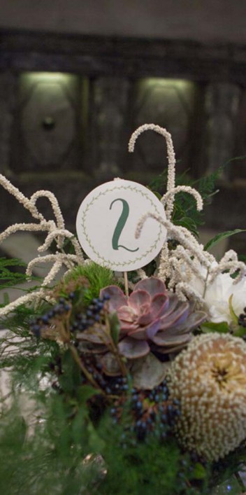 Hochzeitswahn - Ein elegant dekorierter Tisch besteht aus einem Mittelstück mit Sukkulenten und weißen Blumen, spitz zulaufenden weißen Kerzen und einer Tischnummer vor dem Hintergrund eines dunkel gekachelten Raums mit reflektierenden Oberflächen.