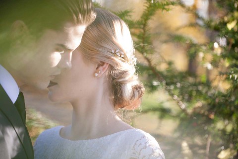 Hochzeitswahn - Ein zärtlicher Moment zwischen einem Paar in Hochzeitskleidung, dessen Gesichter eng beieinander liegen und in sanftes, goldenes Licht gehüllt sind, das durch das umgebende Laubwerk dringt.