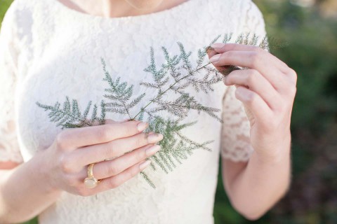 Hochzeitswahn - Eine Frau in einem weißen Spitzenkleid hält zart einen Zweig grünen Farns, Sonnenlicht dringt durch die Blätter und an ihrem Finger ist ein goldener Ring sichtbar.