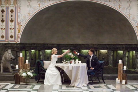 Hochzeitswahn - Ein elegant gekleidetes Paar stößt während eines romantischen Abendessens bei Kerzenschein in einem großen Saal mit aufwendigen Wandgestaltungen, einem großen Kamin und einer majestätischen Wolfsstatue an.
