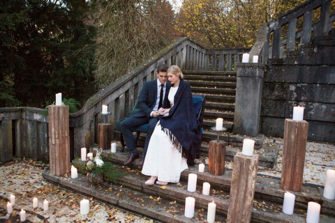 Hochzeitswahn - Ein Paar sitzt eng beisammen auf einer mit brennenden Kerzen geschmückten Holzbrücke und schafft so eine romantische und heitere Atmosphäre inmitten einer rustikalen Herbstkulisse.