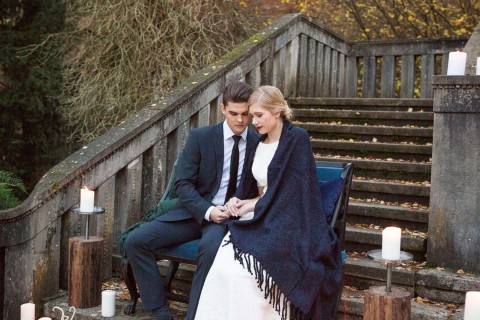 Hochzeitswahn - Ein Paar in formeller Kleidung verbringt einen kontemplativen Moment auf einer Holztreppe, umgeben von brennenden Kerzen, was auf eine intime Zeremonie oder Feier im Freien in der Abenddämmerung schließen lässt.