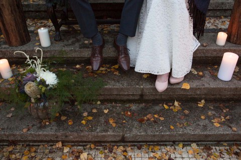 Hochzeitswahn - Ein Paar in formeller Kleidung steht auf Steinstufen inmitten von Herbstblättern. Ihre Intimität wird durch ihre Nähe und die romantische Umgebung angedeutet, die durch Kerzen und ein Blumenarrangement verstärkt wird.
