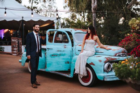 Hochzeitswahn - Ein stilvolles Paar posiert mit einem antiken, türkisfarbenen Pickup-Truck an einem festlichen Veranstaltungsort im Freien und versprüht Vintage-Charme und Feierfreude.