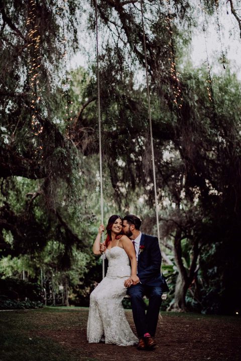 Hochzeitswahn - Ein glückliches Paar teilt einen zärtlichen Moment auf einer Schaukel unter einem mit funkelnden Lichtern geschmückten Blätterdach, das eine romantische, zauberhafte Gartenatmosphäre schafft.