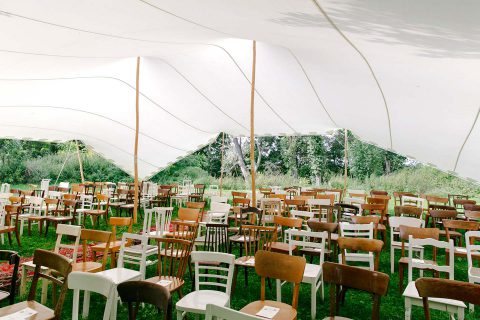 Hochzeitswahn - Ein geräumiges weißes Zelt, das für eine Veranstaltung aufgebaut wurde. Es hat zahlreiche Holzstühle, die auf einen Mittelgang gerichtet sind und von üppigem Grün umgeben sind. Das Zelt ist an den Seiten offen, so dass der Blick auf die natürliche Landschaft draußen freigibt. Wasserburg-Geretzhoven