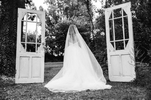 Hochzeitswahn - Eine Braut in einem fließenden weißen Kleid und Schleier steht zwischen zwei freistehenden Vintage-Türen in einer üppigen Waldlandschaft und schafft eine wunderliche und heitere Atmosphäre. Das Bild ist in Schwarzweiß. Wasserburg-Geretzhoven
