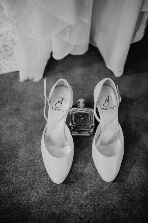 Hochzeitswahn - Ein Schwarzweißfoto von weißen Brautschuhen und einem Parfümflakon auf einem strukturierten Teppich, wobei oben der Saum eines Brautkleides sichtbar ist.