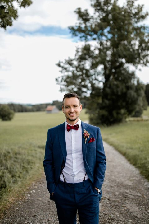 Hochzeitswahn - Ein Mann in einem eleganten blauen Anzug und einer roten Fliege lächelt im Freien auf einem Weg mit grünen Feldern und einem entfernten Haus im Hintergrund.