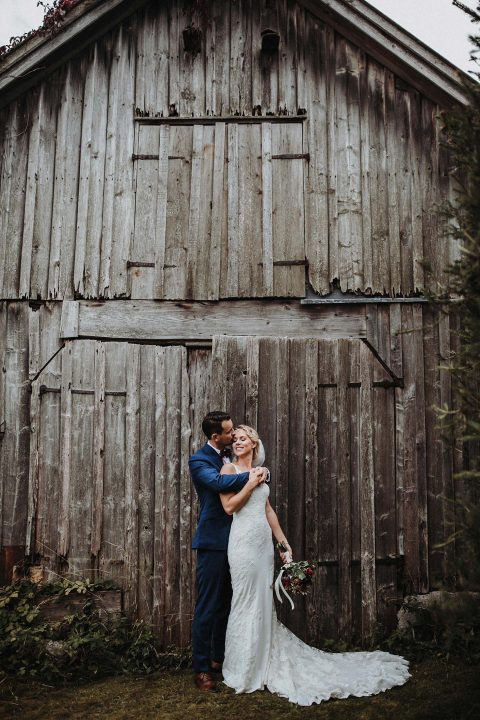 Hochzeitswahn - Ein frisch verheiratetes Paar umarmt sich vor einer rustikalen Holzscheune. Die Braut im weißen Kleid und der Bräutigam im blauen Anzug lächeln freudig. Die verwitterten grauen Bretter der Scheune und die grüne Umgebung verleihen der Szene einen Vintage-Charme.