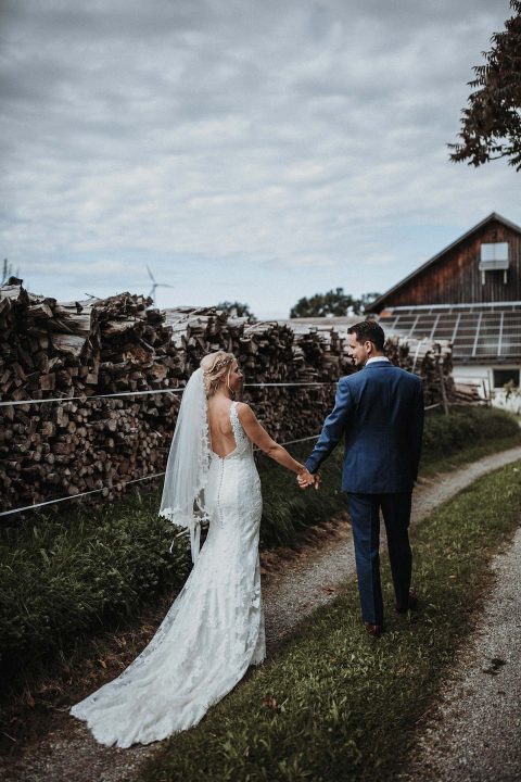 Hochzeitswahn - Eine Braut in einem langen weißen Spitzenkleid und Schleier hält die Hand eines Bräutigams im blauen Anzug, während sie auf einem Landweg neben gestapelten Holzstapeln entlanggehen, im Hintergrund ist eine Scheune zu sehen.