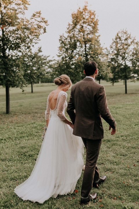 Hochzeitswahn - Braut und Bräutigam gehen Hand in Hand durch einen grünen Park, mit dem Rücken zur Kamera, und strahlen in ihrem gemeinsamen Moment nach der Hochzeitszeremonie ein Gefühl von Intimität und Ruhe aus.