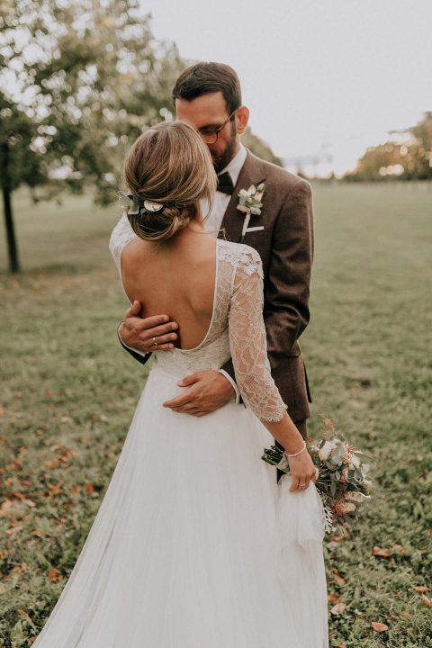 Hochzeitswahn - Eine zärtliche Umarmung zwischen Frischvermählten in einem grünen Park. Die Braut trägt ein Kleid mit Spitzenärmeln und hält einen Blumenstrauß in der Hand. Der Bräutigam trägt einen braunen Anzug als Symbol für Liebe und Einigkeit.