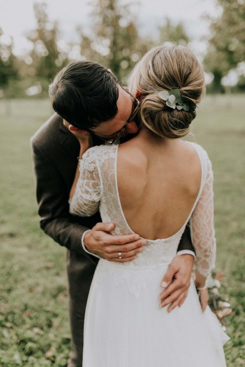 Hochzeitswahn - Ein zärtlicher Moment zwischen einem frisch verheirateten Paar: Der Bräutigam küsst seiner Braut in einer ruhigen Umgebung im Freien liebevoll den Kopf und präsentiert ihr elegantes Spitzenbrautkleid und eine florale Haarnadel.