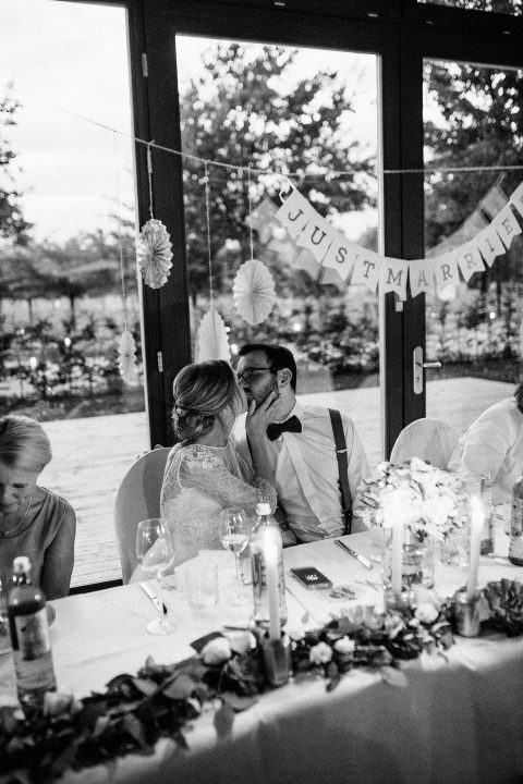 Hochzeitswahn - Ein frisch vermähltes Paar teilt einen zärtlichen Moment bei seinem Empfang. Es sitzt an einem mit Blumen und Kerzen geschmückten Tisch unter einem Banner mit der Aufschrift „Just Married“ (Frisch verheiratet), das ihre Verbindung symbolisiert.